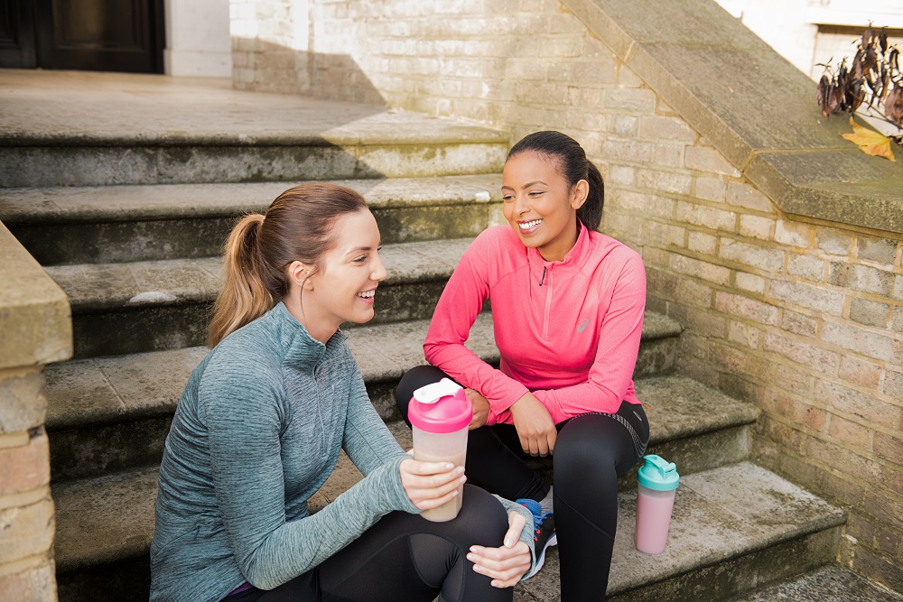 Two ladies sitting on steps outside a house drinking protein shakes