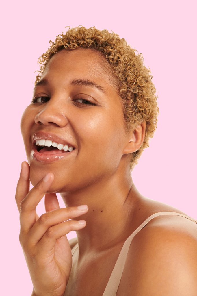 Female laughing with glowing skin and hand on chin with pink background