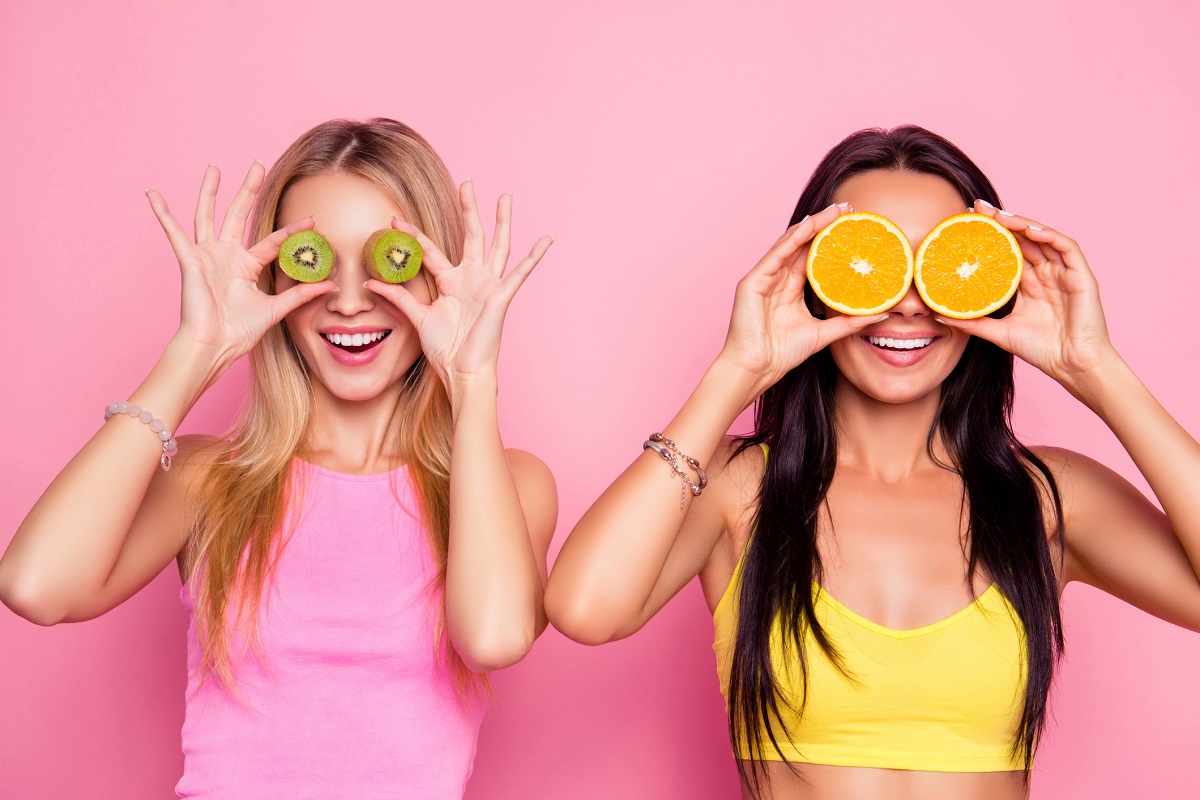 2 women holding fruit in front of their eyes against pink background