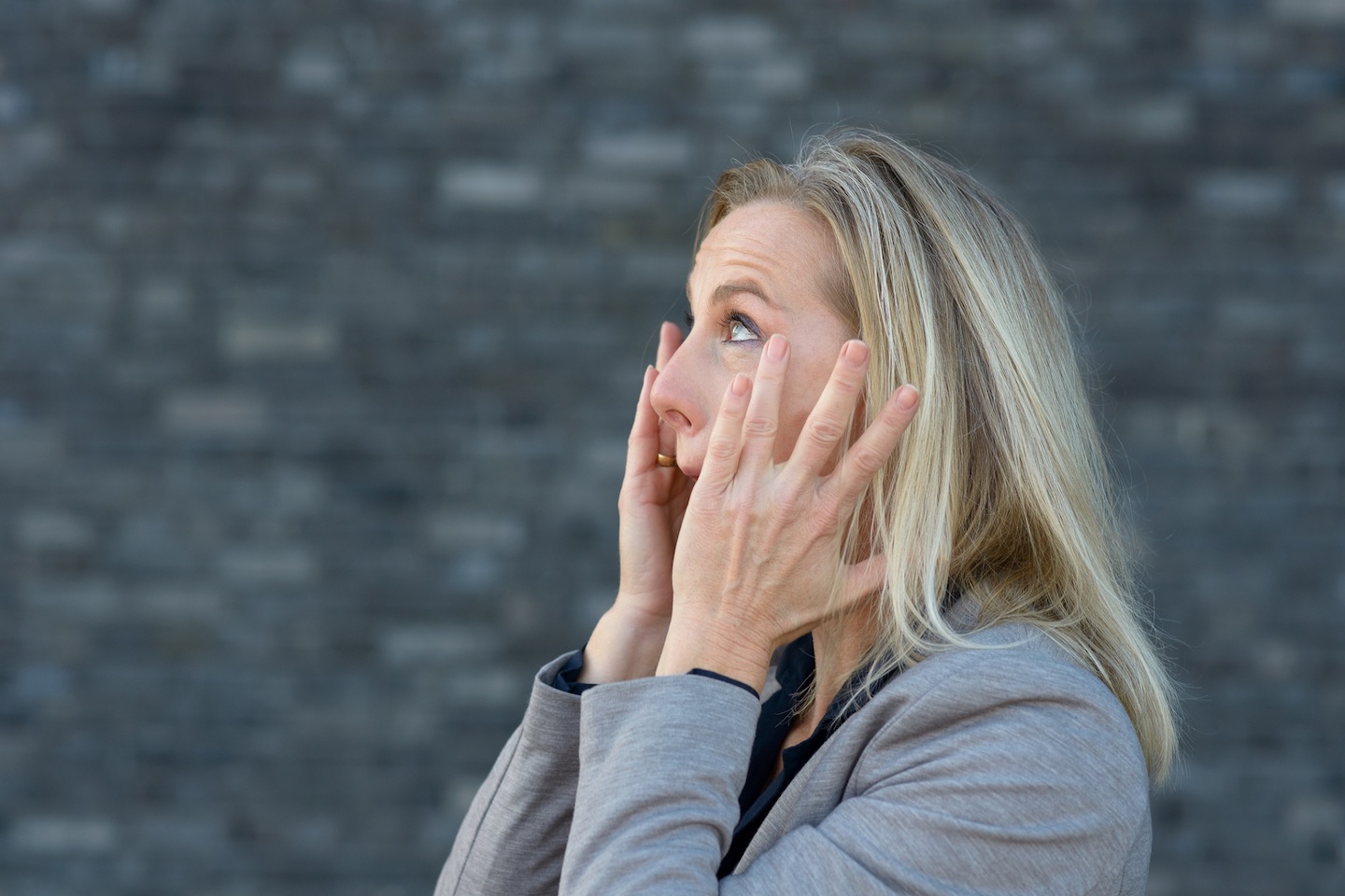 Shocked blond woman with her hands to her cheeks, looking up and screaming. Close-up side portrait against blurred grey background with copy space