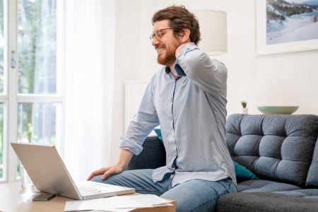 Man sitting on sofa looking down at his laptop, holding his neck in pain