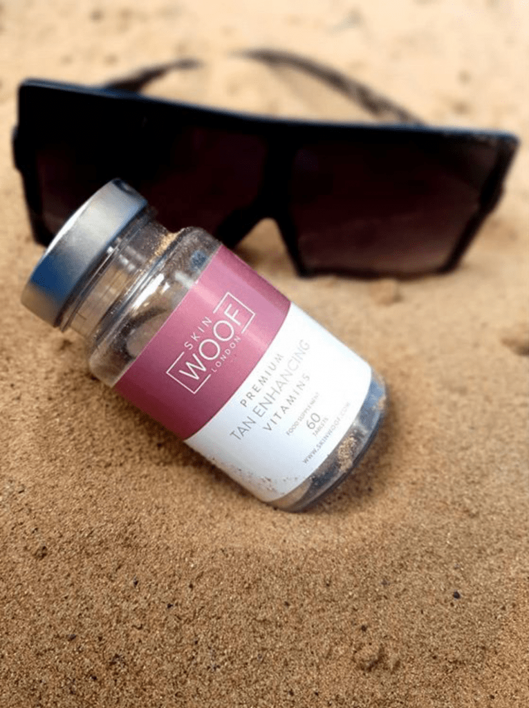 Skin Woof tan enhancing  vitamins bottle in sand with sunglasses in the background